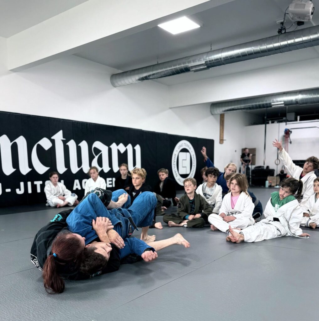 BJJ instructors demonstrating a ground technique to a seated youth class at Sanctuary Jiu-Jitsu Academy in Madison.