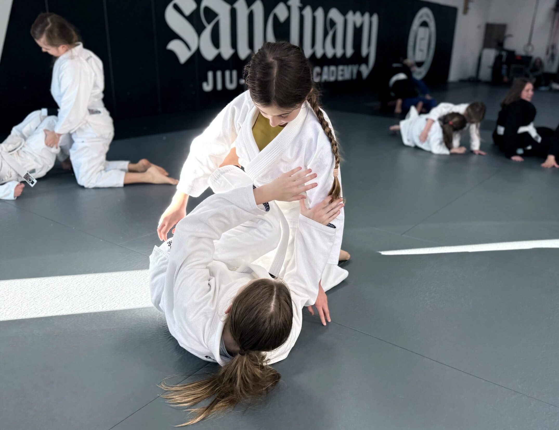 Girls jiu-jitsu program students in white gis drilling a technique together at Sanctuary Jiu-Jitsu Academy.